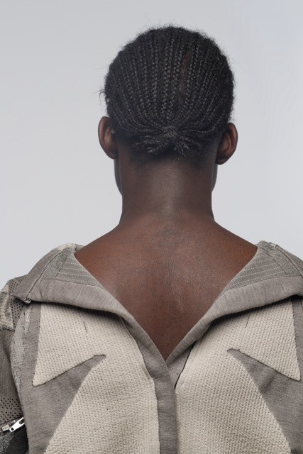 Rear view of a person with intricately braided hair facing away. They wear a textured garment made of Merino wool, showcasing a striking geometric design on the back. The background remains a neutral, light color.