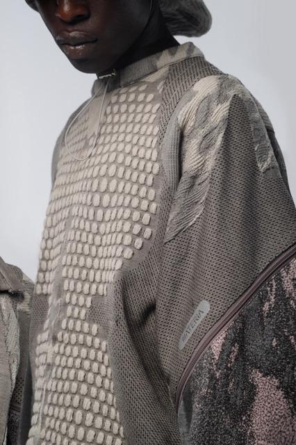 A person wearing a textured, multi-patterned gray sweater crafted from Merino wool with unique mesh and patch designs. The sweater has a mix of geometric and abstract textures. The person is partially visible, focusing on their attire.
