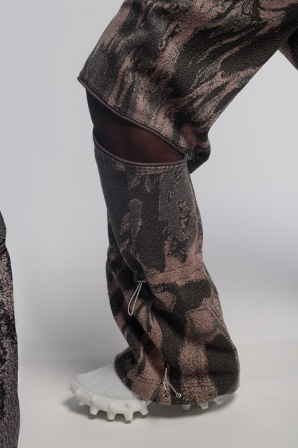 A person wearing patterned, textured pants crafted from Merino wool showcases a rough design. The lower part features zippered sections and white, rugged shoes with textured soles. The plain background accentuates the intricate clothing details.