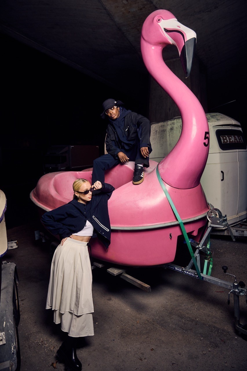 A man dressed in blue including a wool hoodie and a long wool jacket which has a Südwolle Group logo on it and wool trousers. He is leaning against a woman with a white wool skirt and a blue wool jacket. Both their styles are very Urban Activewear. He is sittingon a boat which is shaped like a flamingo. The woman is standing on the ground and leans against the boat