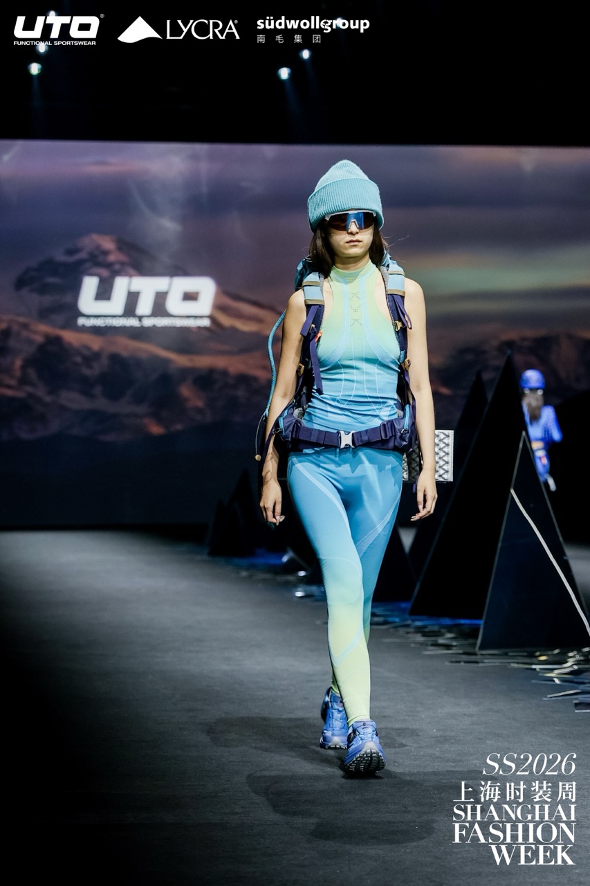 A model walks the runway wearing a blue and yellow sporty outfit with a backpack, blue beanie, and sunglasses at Shanghai Fashion Week SS2026. The backdrop features a mountain landscape and event sponsor logos.