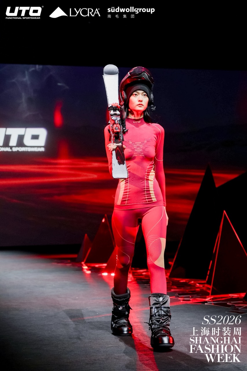 A female model in a red patterned bodysuit, black snow boots, and a helmet walks the runway holding skis at Shanghai Fashion Week SS2026, with sponsor logos in the background.