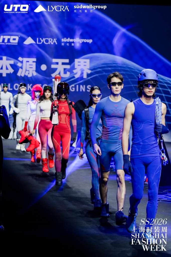 Models wearing futuristic, form-fitting athletic wear in shades of blue, red, and purple walk down the runway at Shanghai Fashion Week, with a blue-lit abstract background and brand logos visible behind them.