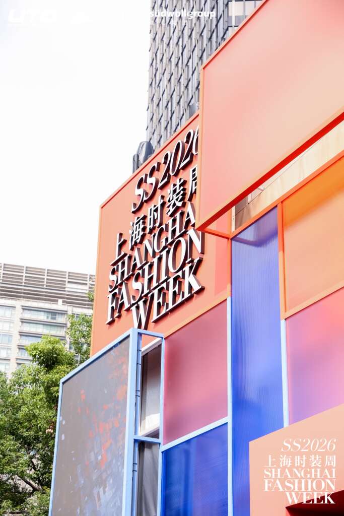 A large, colorful outdoor sign reads SS2026 Shanghai Fashion Week in English and Chinese, set against tall city buildings and trees. The modern sign features orange, blue, and pink panels.