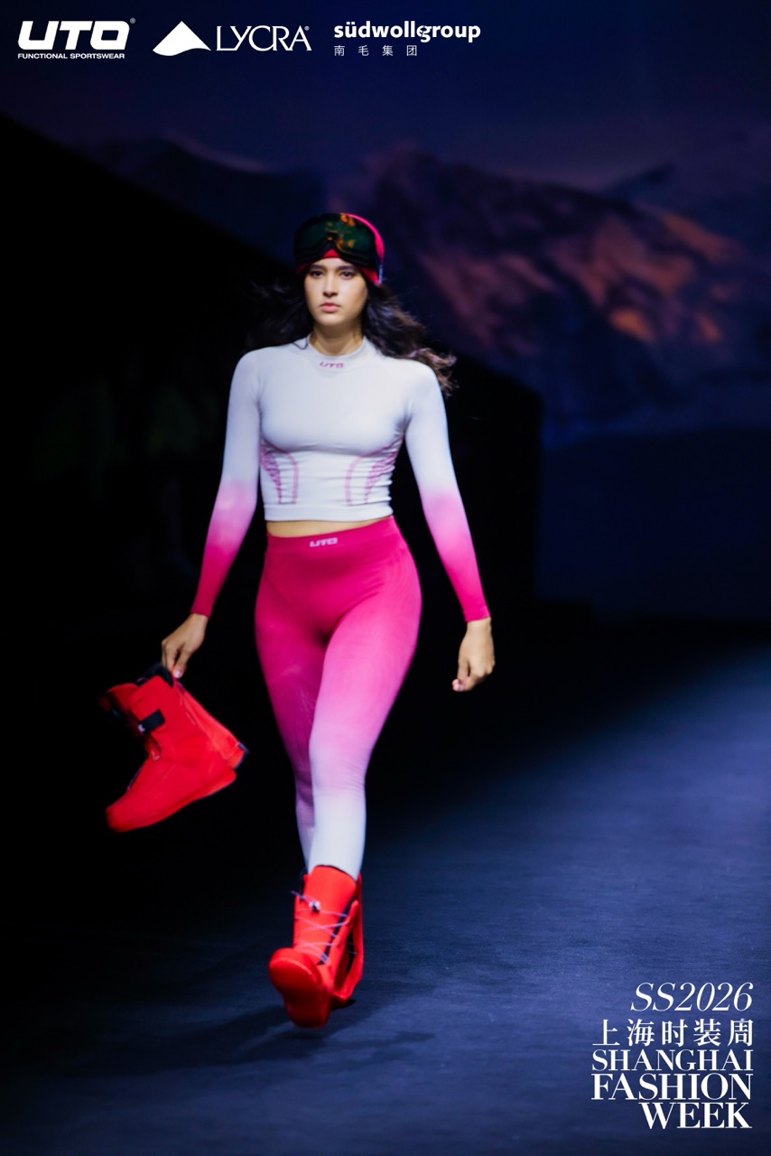 A model walks the runway at Shanghai Fashion Week in a gradient pink and white fitted outfit with red boots and a red headband. The background features mountain imagery and event sponsor logos.