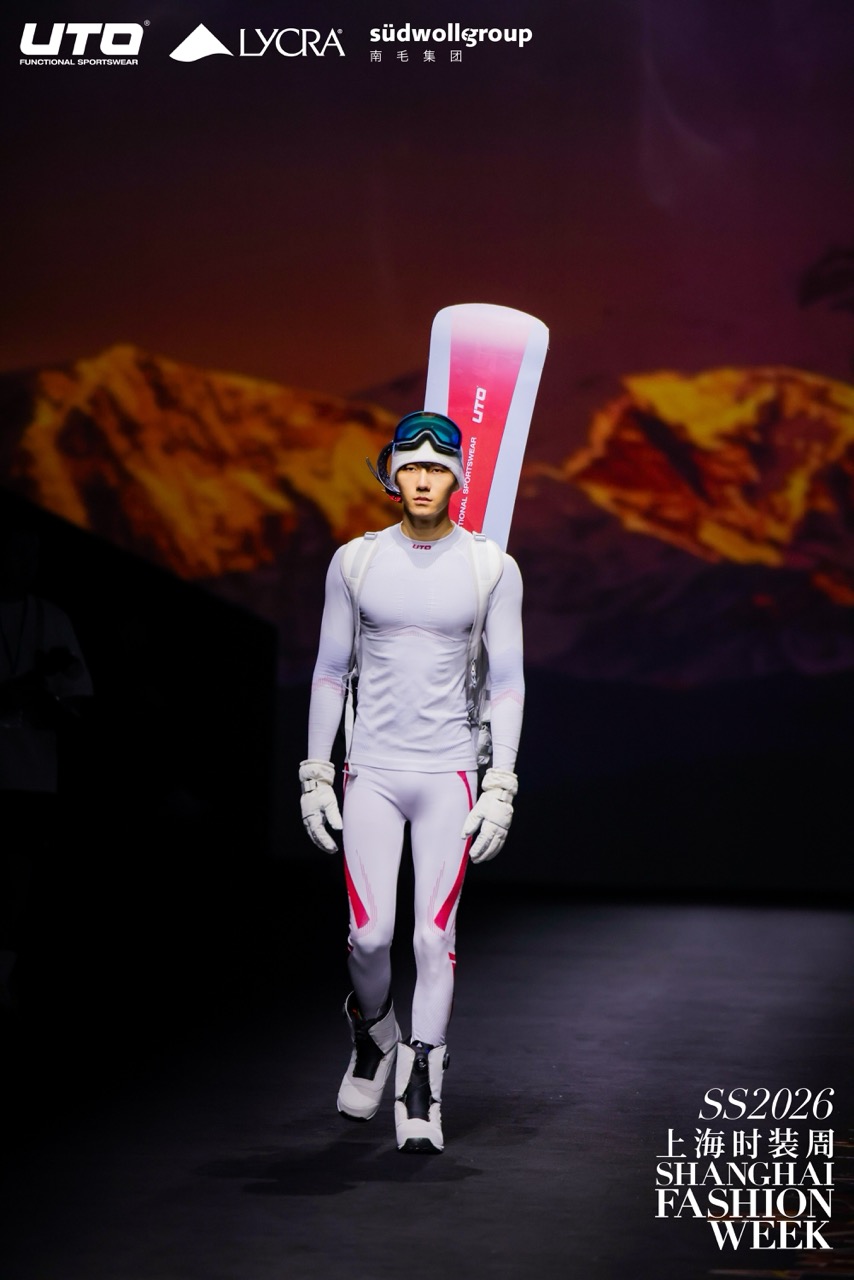 A male model in a white and pink ski outfit with goggles and a helmet walks the runway carrying skis at Shanghai Fashion Week SS2026. Bright mountain imagery forms the backdrop. Fashion brand logos are at the top of the image.