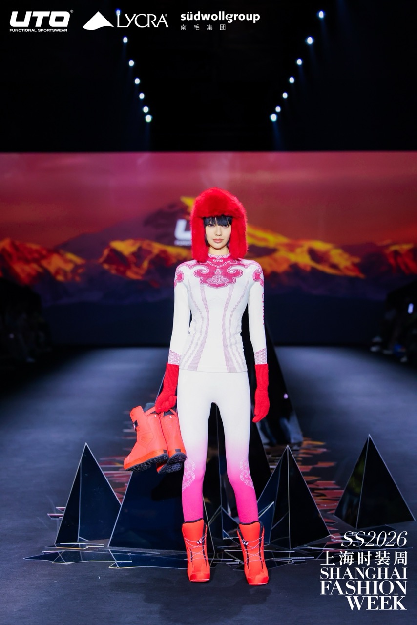A model wearing a white and pink bodysuit, red fur hat, and red boots stands on a runway holding matching boots. The backdrop displays a mountain landscape. Text reads SHANGHAI FASHION WEEK SS2026.