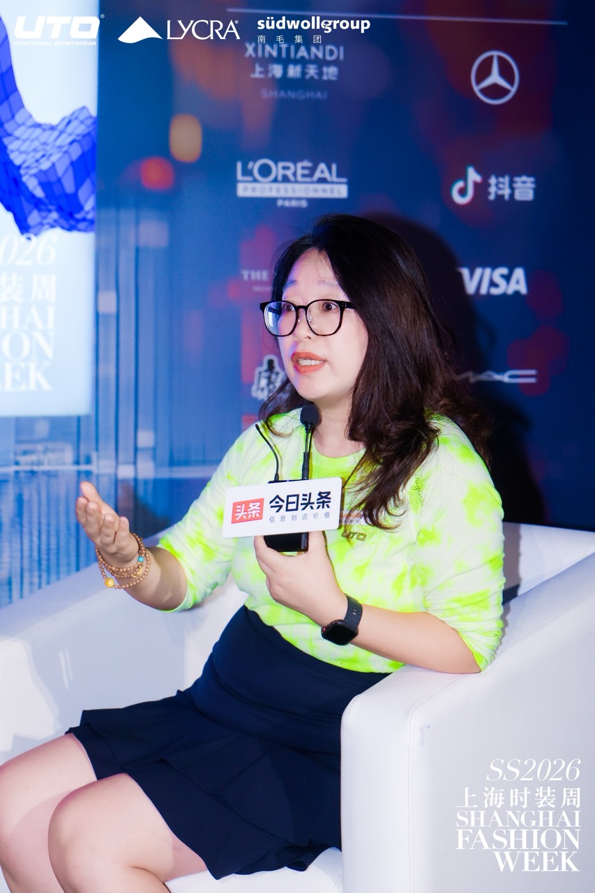 A woman with long hair, glasses, and a neon green shirt sits in a white armchair, speaking into a microphone at Shanghai Fashion Week 2026. Event sponsor logos are visible on a blue background behind her.