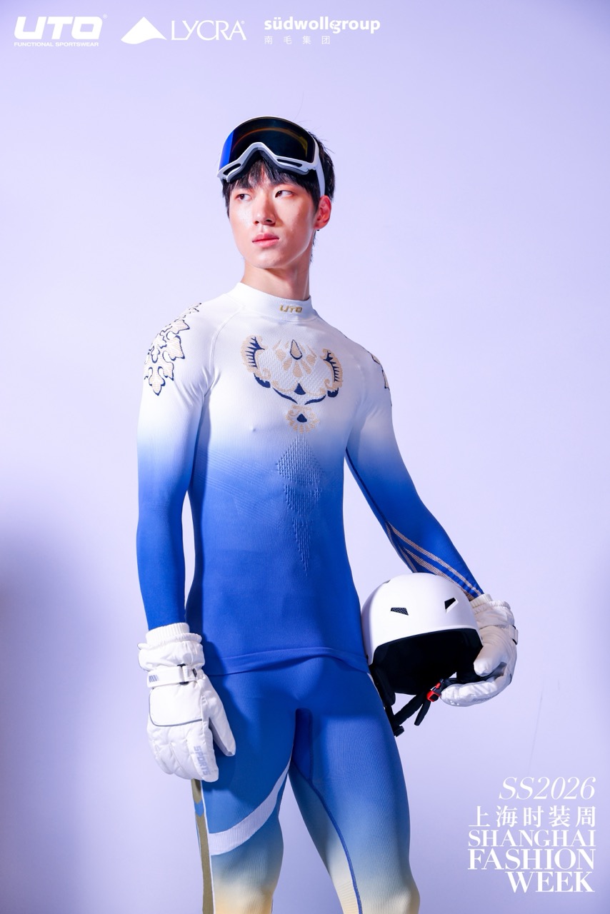A male model in a blue and white gradient ski suit holds a white helmet and ski goggles. He stands against a light background with Shanghai Fashion Week SS2026 and sponsor logos visible.