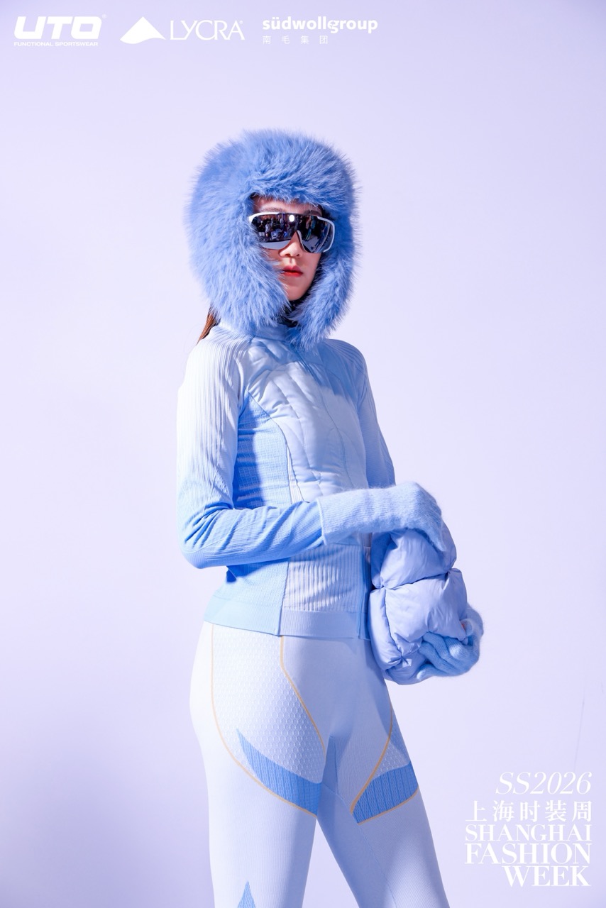 A model in a blue fur hood, matching gloves, sporty fitted jacket, and leggings poses confidently against a pale background. She wears large sunglasses and holds a puffy blue accessory. Shanghai Fashion Week SS2026 text appears at the bottom.