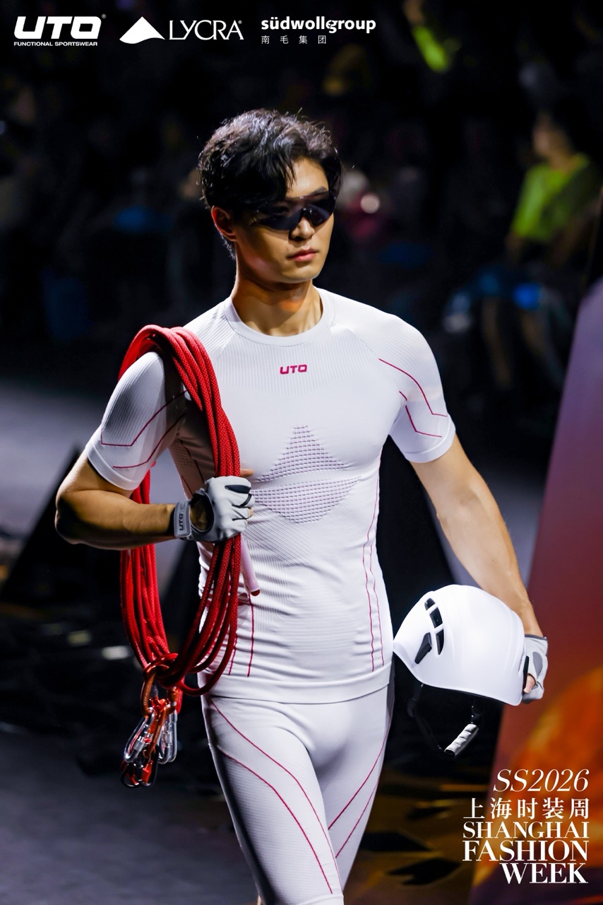 A male model in a white, form-fitting athletic outfit with red accents walks the runway at Shanghai Fashion Week. He wears sunglasses, carries climbing rope over one shoulder, and holds a white helmet in his hand.
