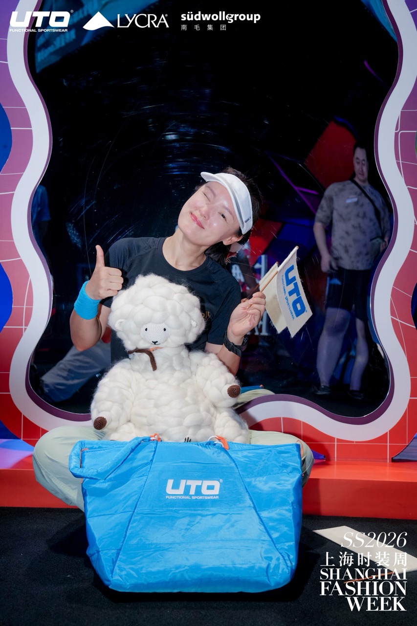 A smiling person in a cap and UTO T-shirt poses with a thumbs up, holding a UTO flag, sitting behind a fluffy sheep doll and a blue UTO bag at Shanghai Fashion Week SS2026.