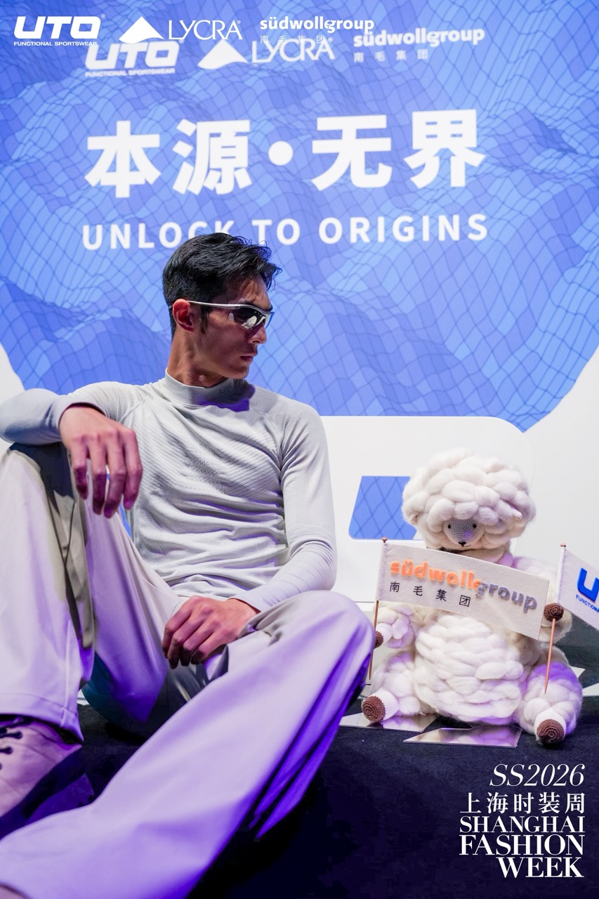 A male model in sunglasses and a cream outfit poses next to a plush sheep holding a südwollgroup flag. Behind them are blue logos and text promoting Unlock to Origins at Shanghai Fashion Week SS2026.
