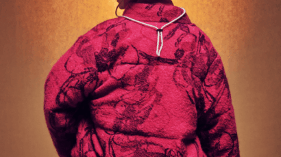 Person facing away, wearing a textured, oversized magenta jacket with dark patterns, a tan bucket hat, and a necklace, standing against a warm, gradient brown background.