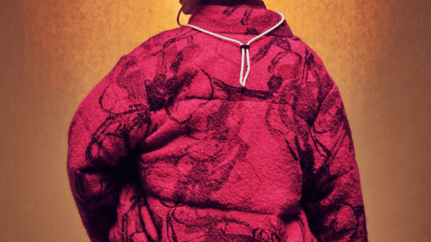 Person facing away, wearing a textured, oversized magenta jacket with dark patterns, a tan bucket hat, and a necklace, standing against a warm, gradient brown background.