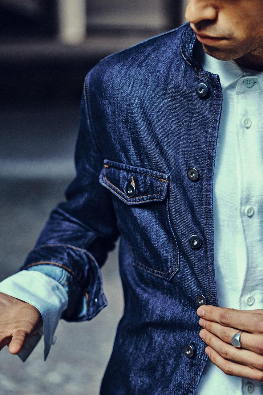 A man wearing a blue denim jacket over a white button-up shirt, adjusting his jacket with one hand. Only his torso and part of his face are visible.