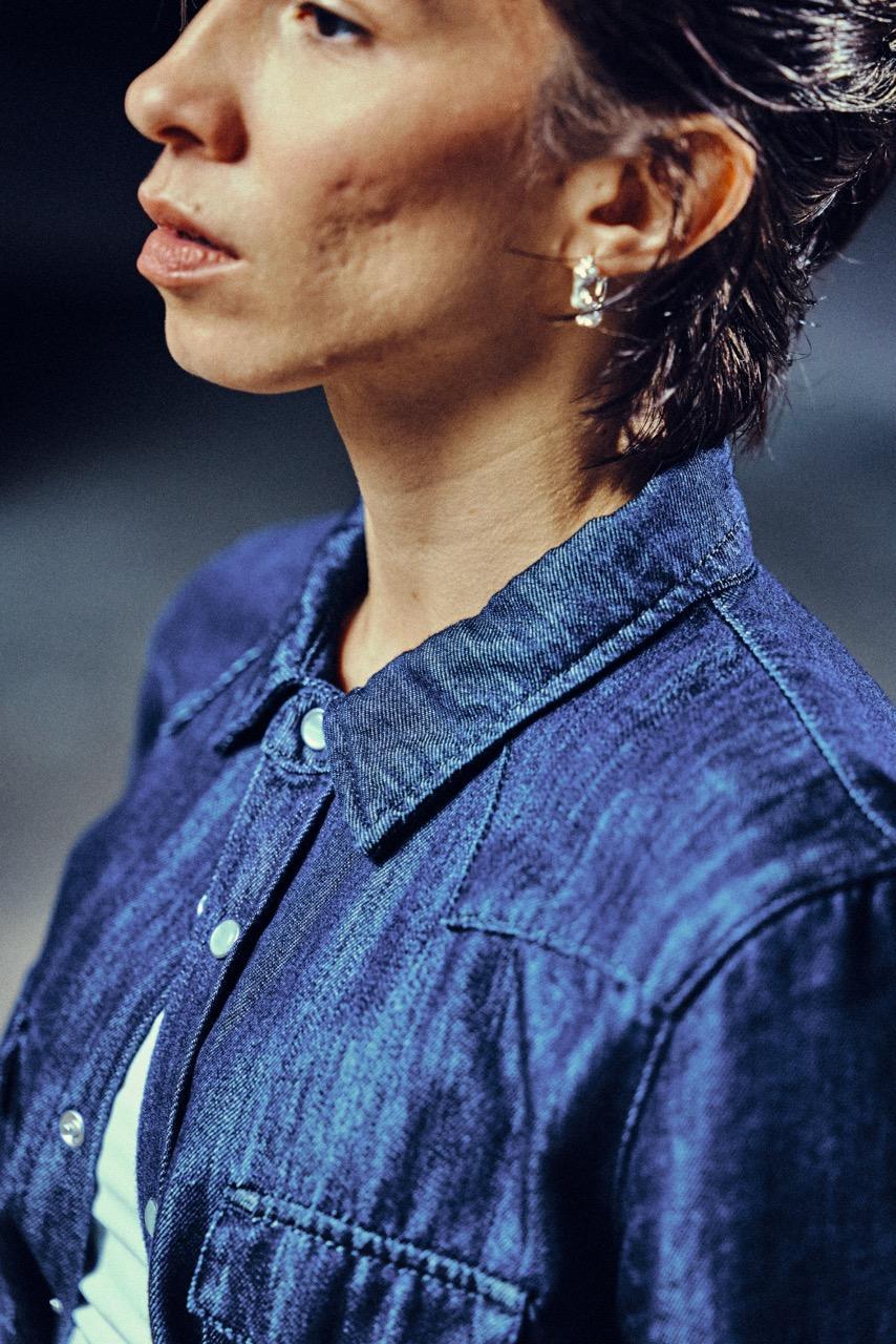 A person with short brown hair wearing a blue denim button-up shirt and small hoop earrings is shown in a close-up, angled view. The background is blurred, focusing attention on the face and upper body.