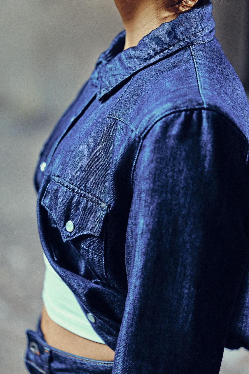 A person wearing a dark blue denim jacket over a white top, shown from the chest up, with the background out of focus.