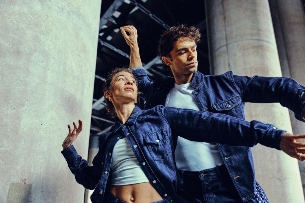 Two people wearing denim outfits pose gracefully with outstretched arms, standing close together in front of large concrete pillars. They appear focused, as if dancing or performing under an industrial structure.