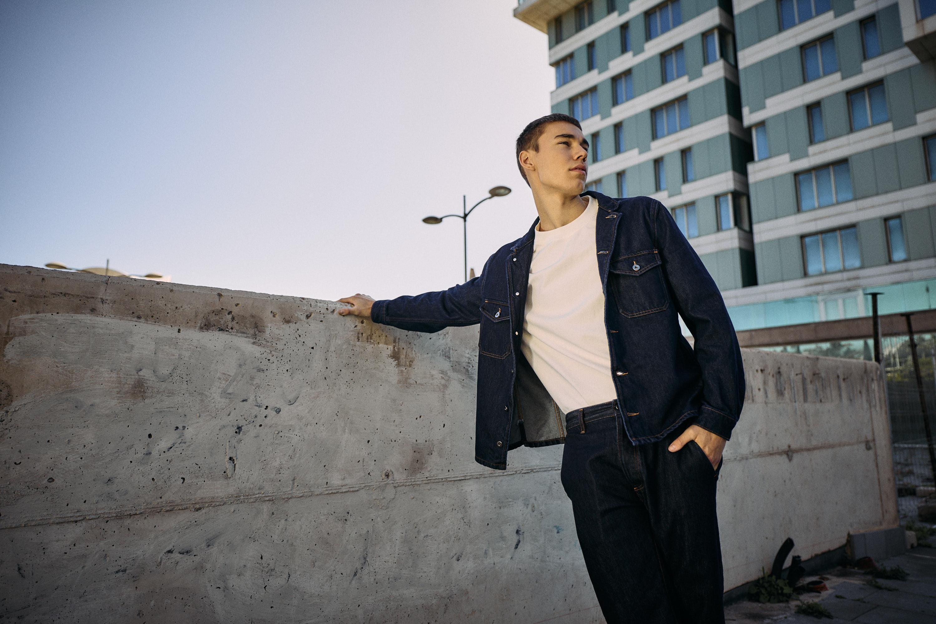 A young man in a white shirt and dark denim jacket leans against a concrete wall outdoors, with a multi-story building and clear sky in the background.