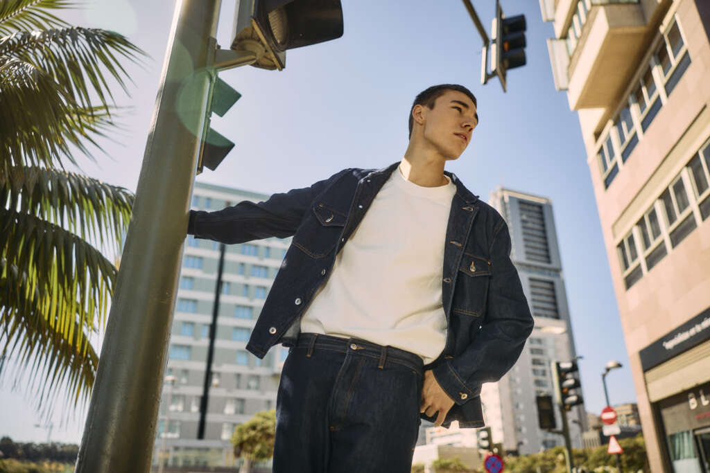 A young man in a denim jacket and jeans stands on a city street corner, holding onto a traffic pole, with tall buildings, palm trees, and traffic lights visible under a clear blue sky.
