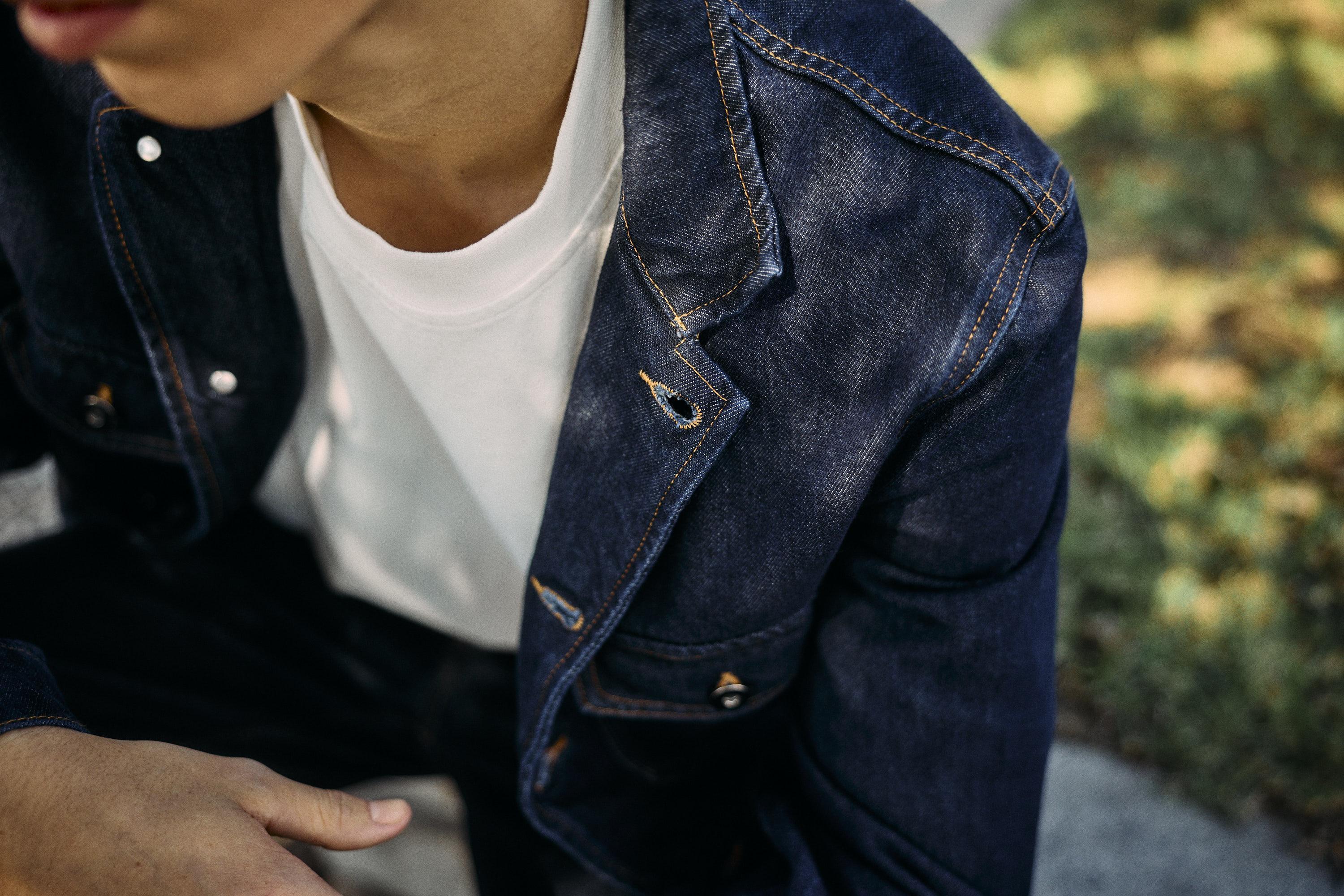 A person wearing a white t-shirt under a dark blue denim jacket sits outdoors on a sunny day, with green grass and a sidewalk visible in the background. The persons face is partially out of frame.