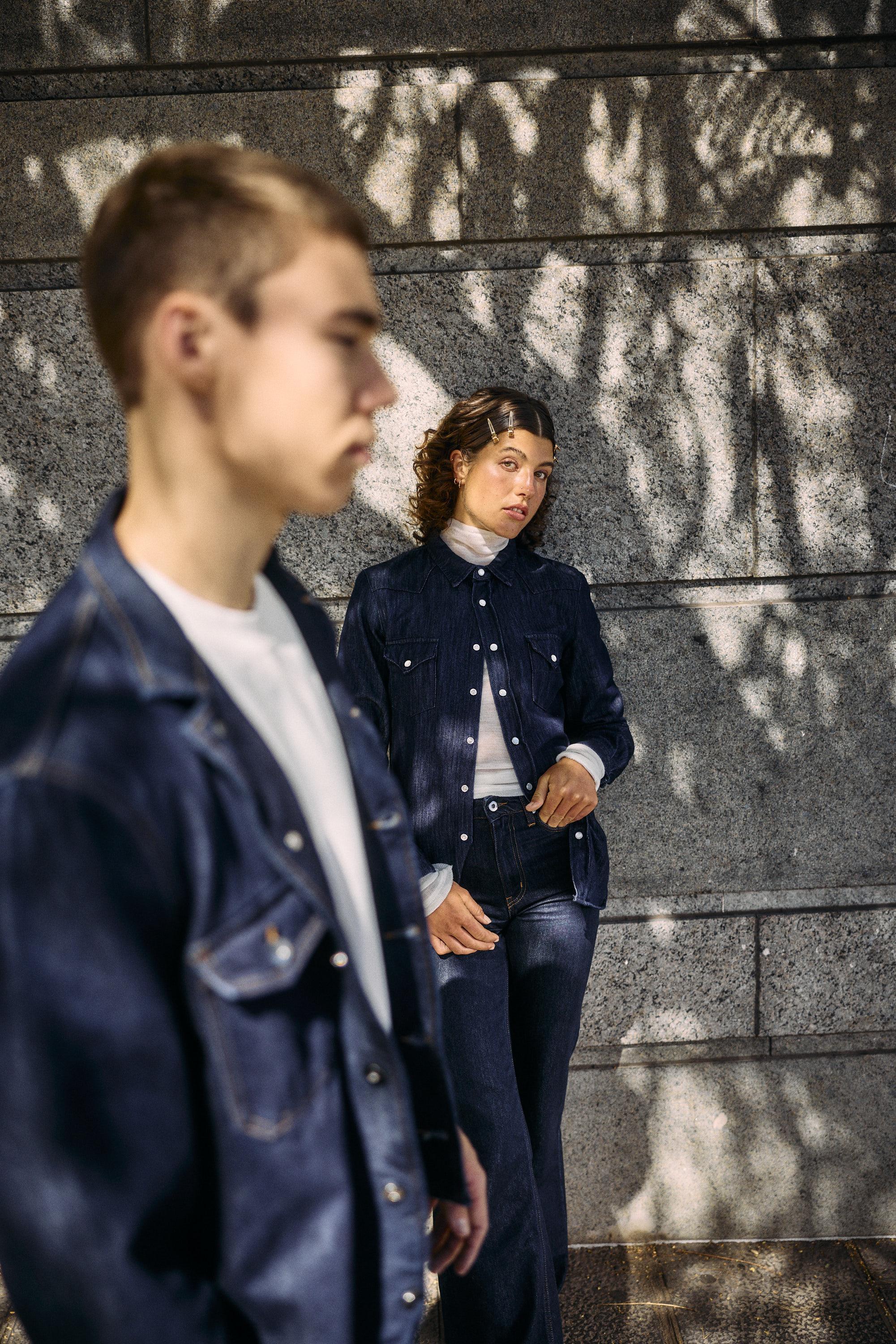 A young woman in denim clothing stands against a sunlit stone wall with dappled shadows, while a young man in a denim jacket stands in the foreground, slightly out of focus.