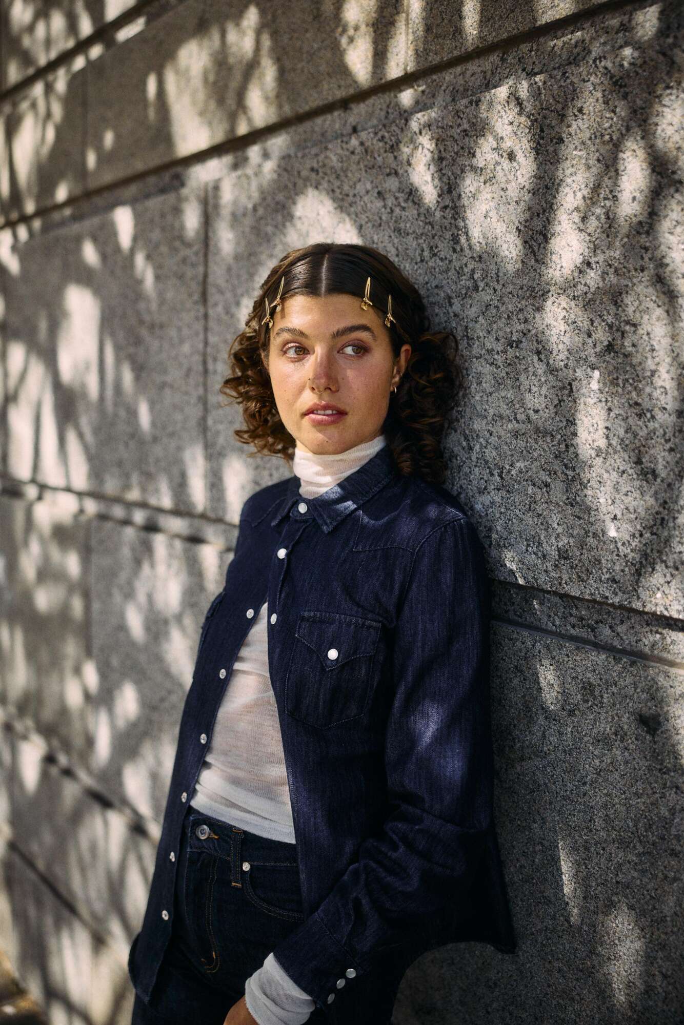 A young woman with wavy hair, wearing gold hair clips, a white turtleneck, and a dark denim jacket, stands against a sunlit stone wall with dappled shadows from nearby trees. She looks thoughtfully into the distance.
