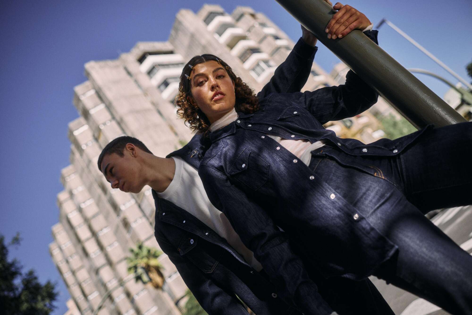 Two people wearing denim jackets and jeans pose confidently outdoors in front of a tall, modern apartment building under a clear blue sky. One leans on a pole, while the other looks away.