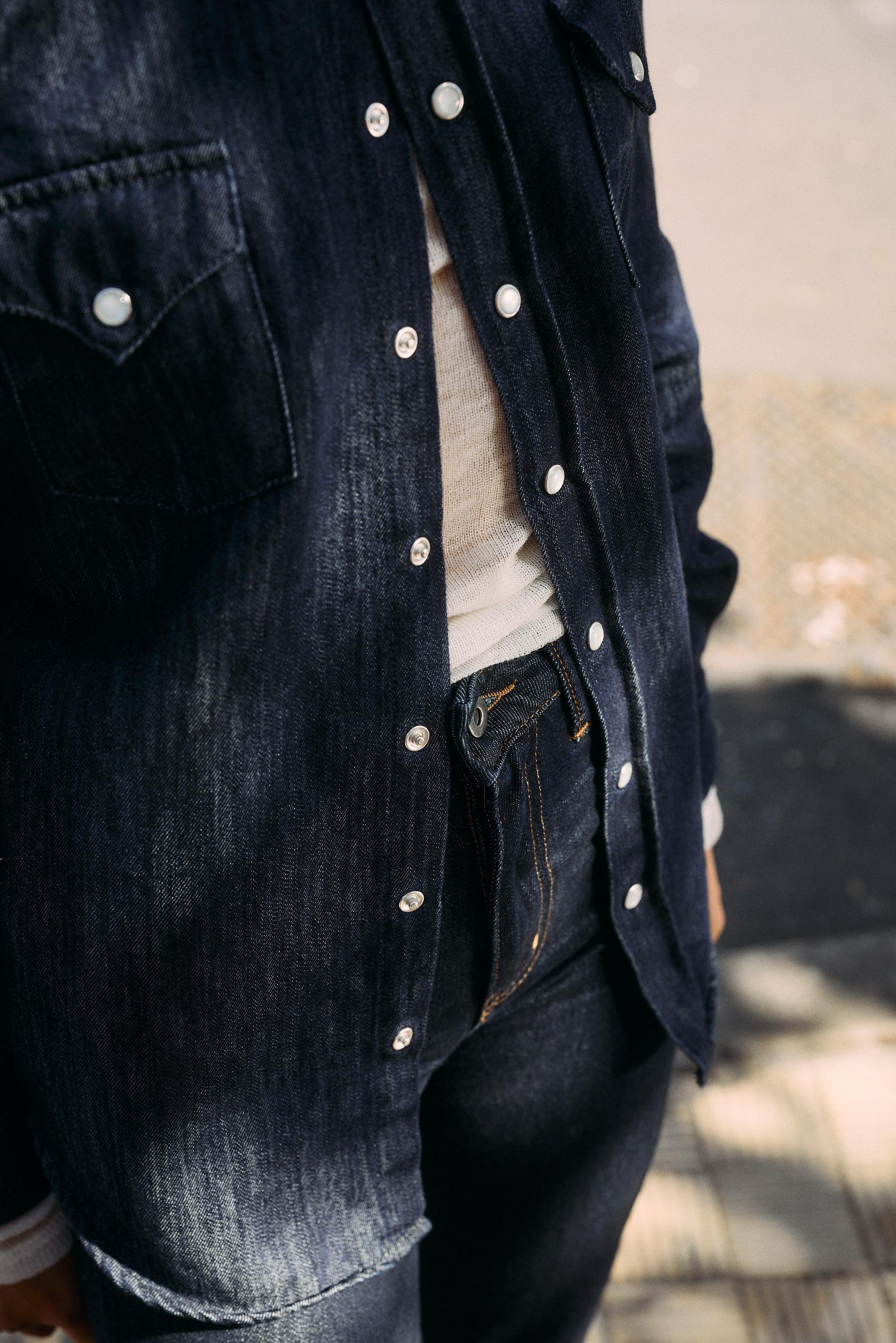 A person wearing a dark denim shirt over a light-colored top and matching dark denim jeans stands outdoors, with pavement and a metal grate visible in the background.