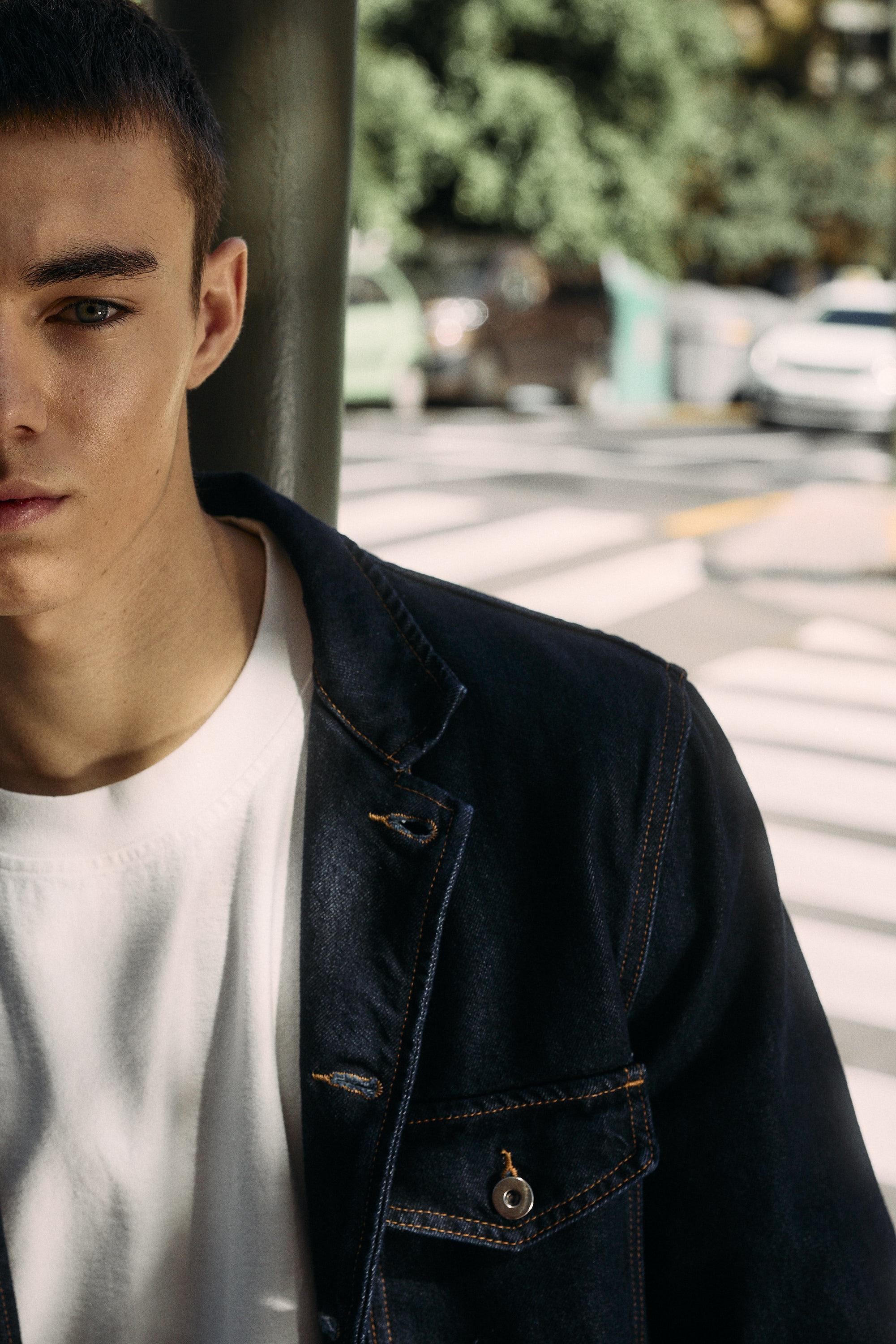 A young man wearing a dark denim jacket over a white shirt stands outdoors near a crosswalk, with trees and parked cars visible in the blurred background. Only half of his face and upper body are shown.