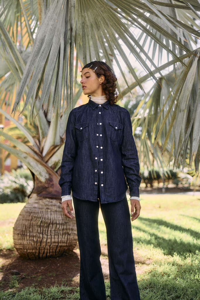 A person stands outdoors under a large palm tree, wearing a dark denim button-up shirt over a light turtleneck and matching dark denim pants, looking to the side with hands by their sides. Sunlight filters through the palm fronds.