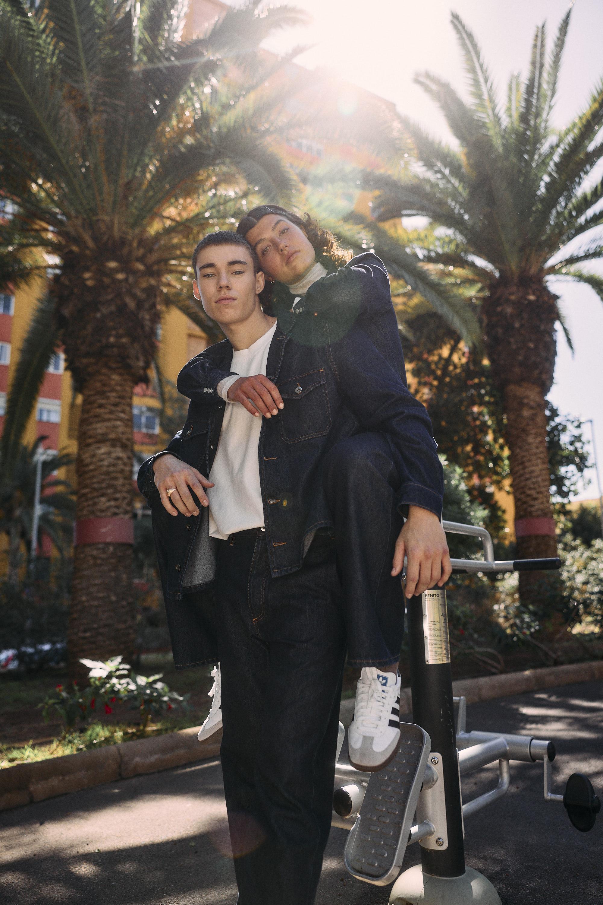 Two young people pose confidently outdoors among palm trees. One stands in front wearing a dark denim jacket and white shirt, while the other leans on their shoulder, dressed in denim and white sneakers. Sunlight streams through the trees.