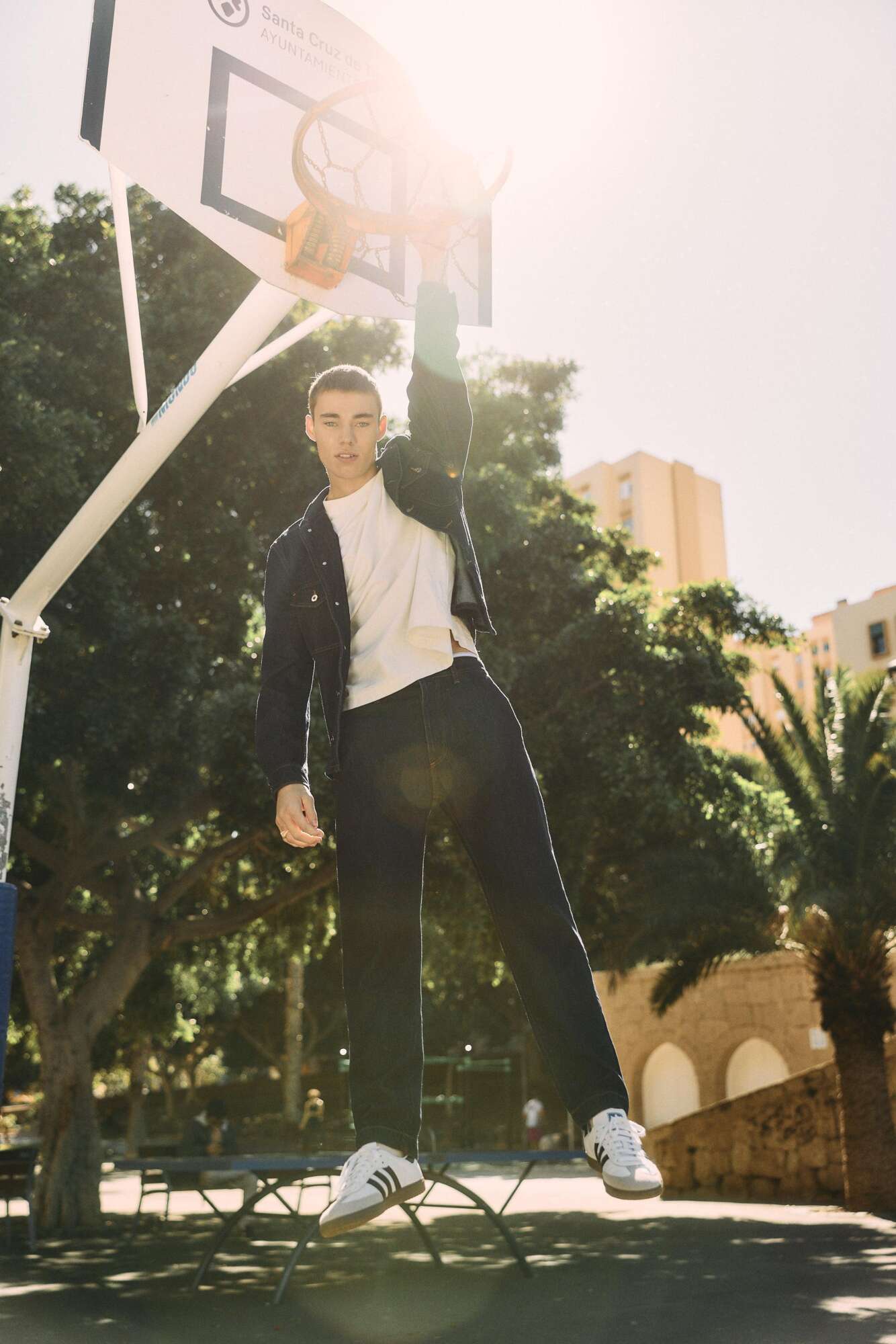 A young man hangs from a basketball hoop with one hand on an outdoor court, wearing a white shirt, dark jacket, and pants, with sunlight shining through trees and buildings in the background.