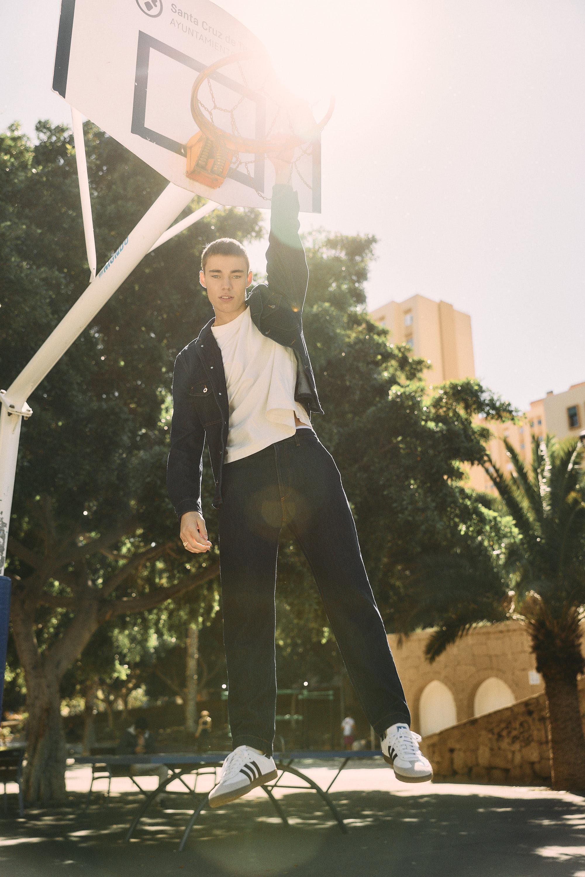 A young man hangs from a basketball hoop with one hand on an outdoor court, wearing a white shirt, dark jacket, and pants, with sunlight shining through trees and buildings in the background.