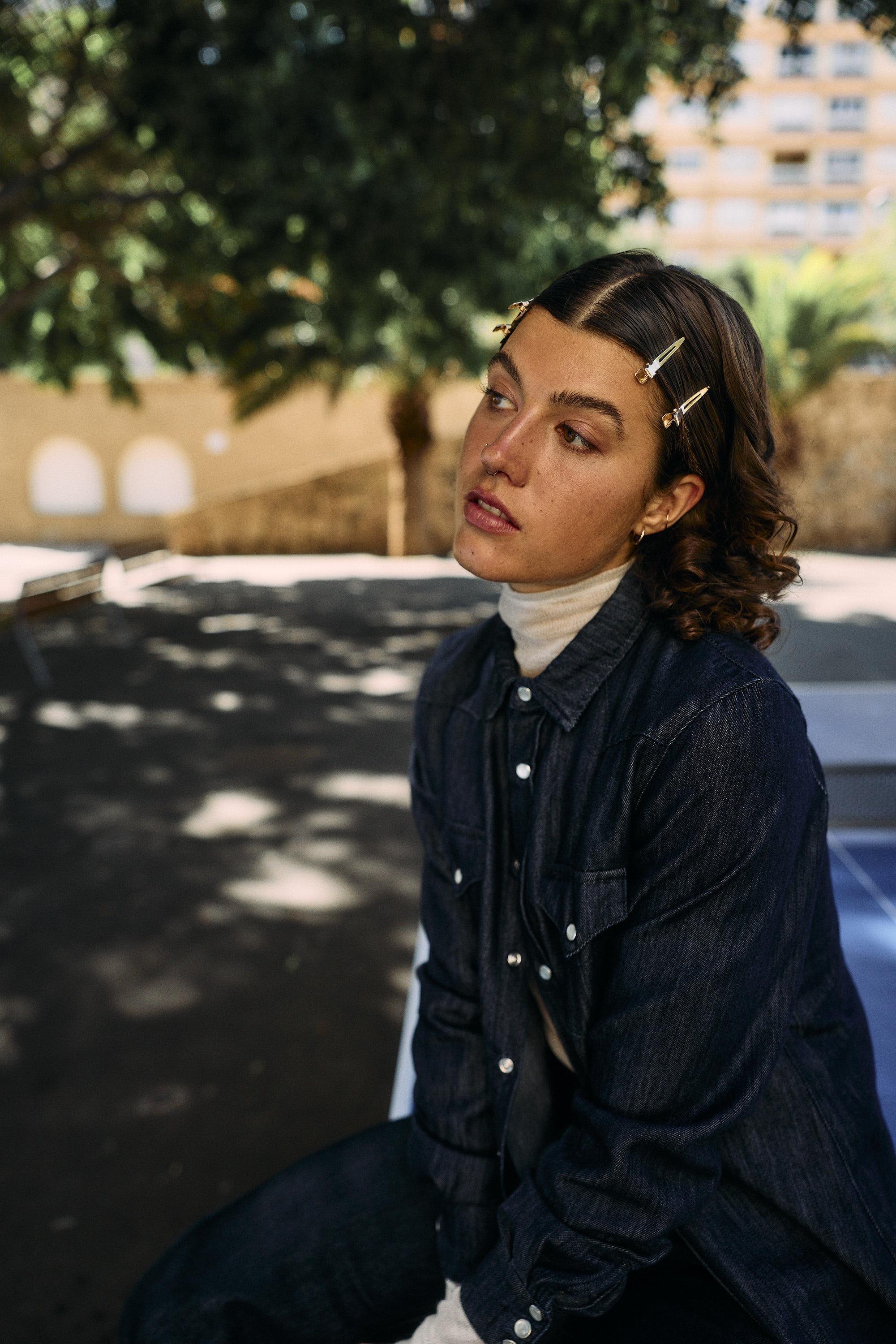 A woman with wavy brown hair pinned back by several clips sits outdoors, wearing a dark denim shirt over a white turtleneck. She looks thoughtful, with trees and a blurred building in the background.