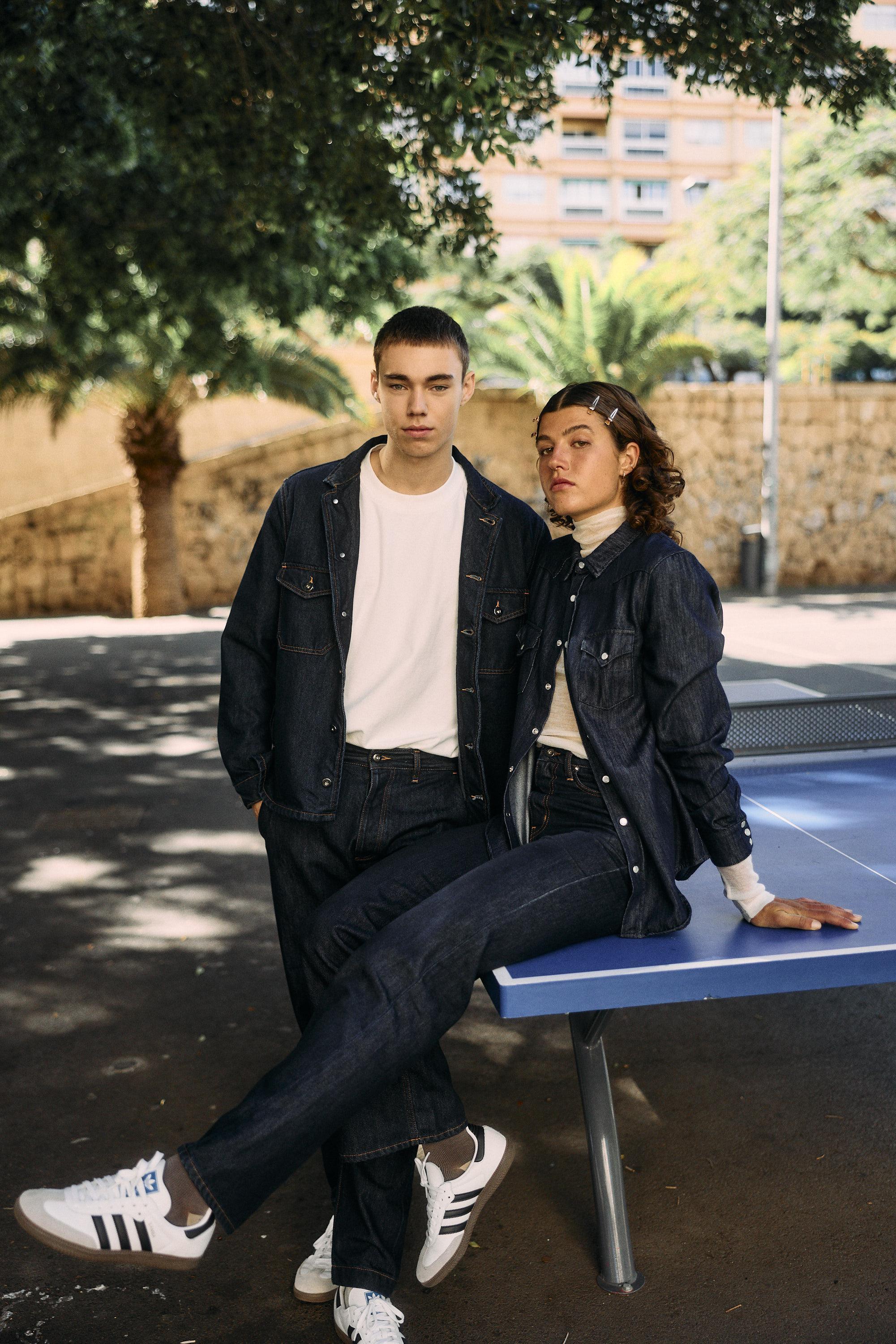 Two people in dark denim outfits and white shirts pose by a blue outdoor ping pong table. One sits on the table with legs crossed; the other stands beside them. Trees and a brick wall are in the background.