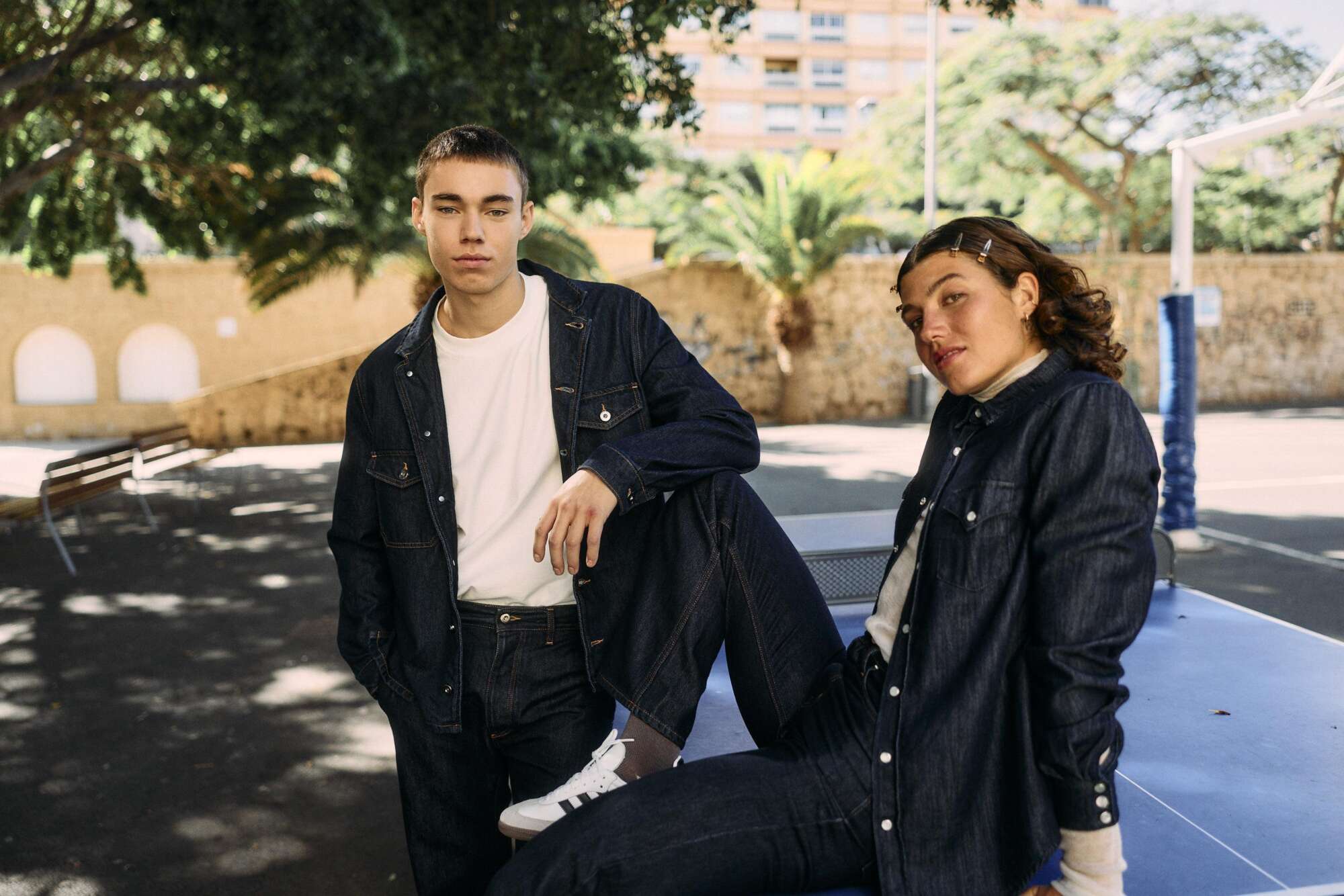 Two young people pose outdoors in matching dark denim outfits. One stands with a neutral expression and the other sits with a leg up on a bench, leaning casually. Trees and a building are visible in the background.