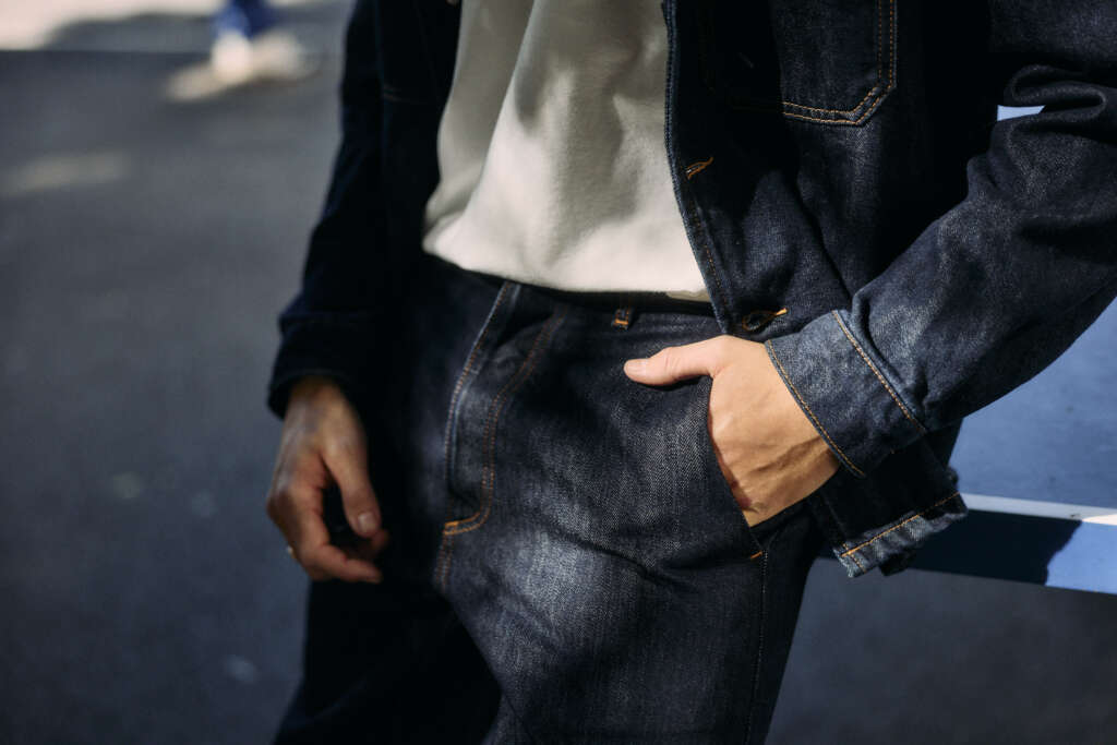 A person wearing a white shirt and dark denim jacket and jeans stands outdoors with one hand in their pocket, leaning casually against a blue surface.