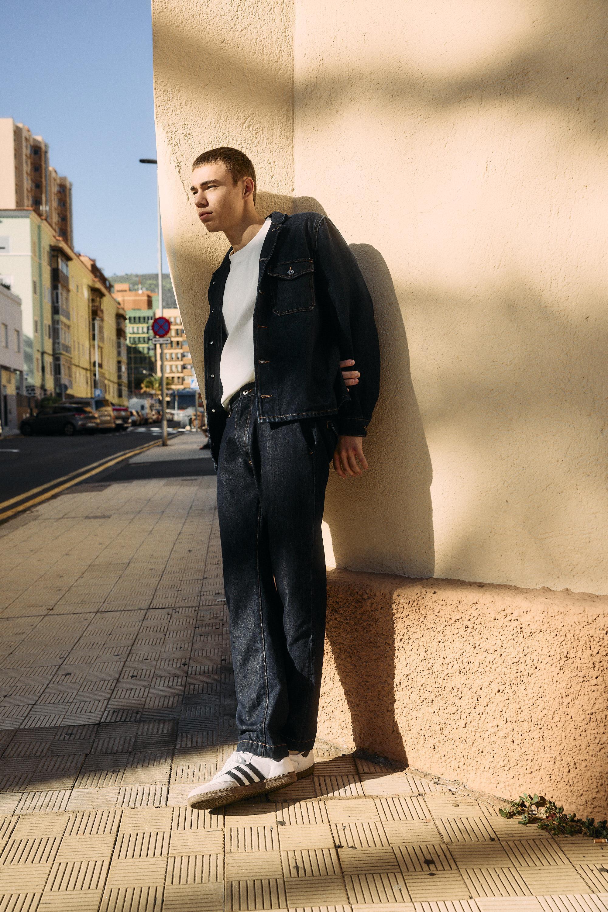 A man wearing a dark denim jacket and jeans, a white T-shirt, and white Adidas sneakers stands leaning against a beige wall on a sunlit city street. Shadows and buildings line the background.
