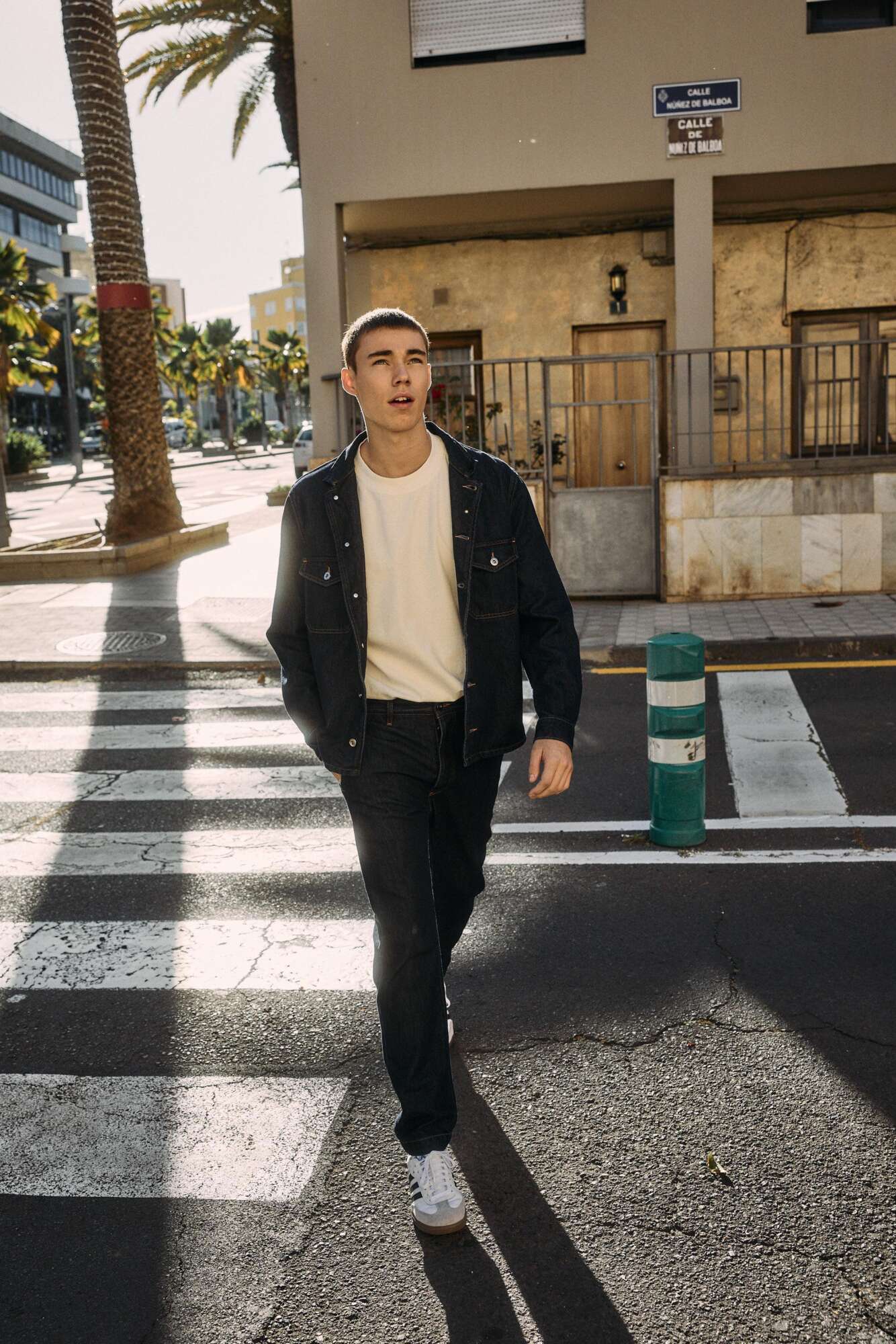 A young man wearing a denim jacket, black jeans, and white sneakers walks across a sunlit crosswalk on a city street, with palm trees and buildings in the background.