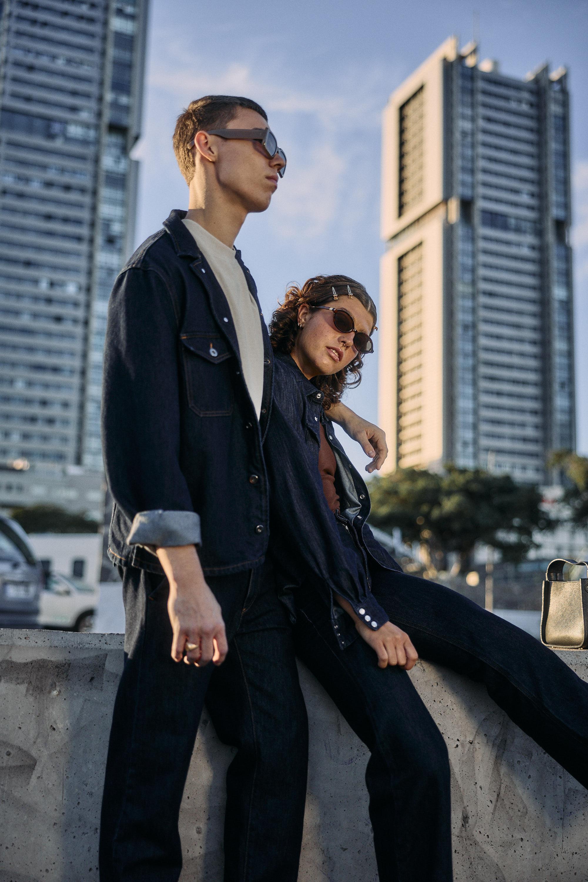 A man and woman wearing sunglasses and dark denim outfits lean against a concrete barrier with tall modern buildings and blue sky in the background. The woman sits, relaxed, while the man stands beside her.