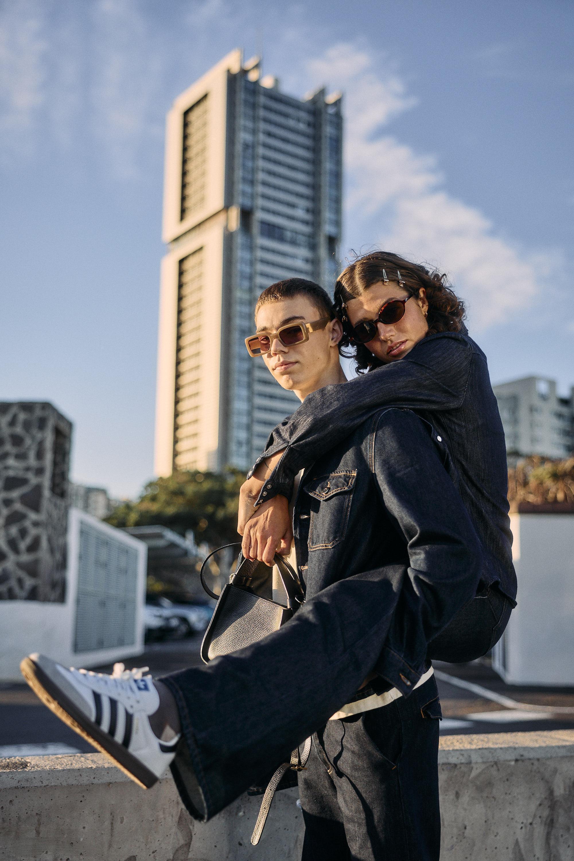 Two people wearing sunglasses and denim clothing pose together outdoors. One person stands while the other wraps their arms around their shoulders, with legs lifted. A tall modern building and a blue sky are visible in the background.
