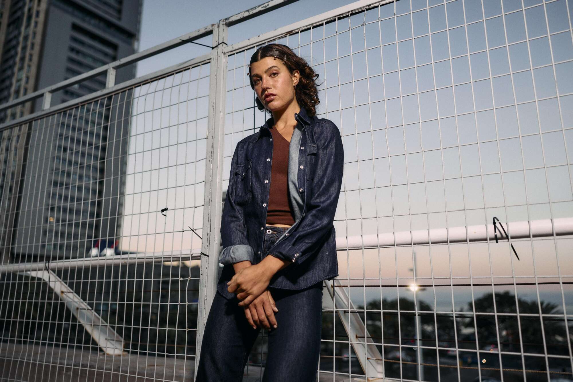 A person with curly hair stands in front of a metal fence at dusk, wearing a denim jacket and jeans, with city buildings and a soft sunset sky in the background.