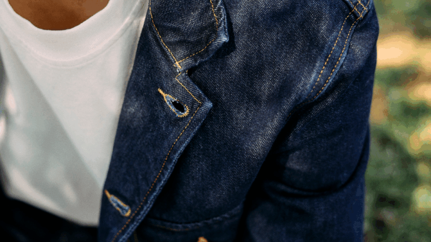 A person wearing a white t-shirt and a blue denim jacket is seen from the shoulders down. A golden New ribbon appears in the top left corner. The background is blurred greenery and pavement.