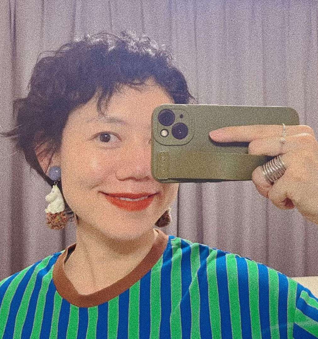 Person with short curly hair takes a mirror selfie, smiling, wearing a blue and green striped shirt, statement earrings, and rings. Beige curtains are in the background.
