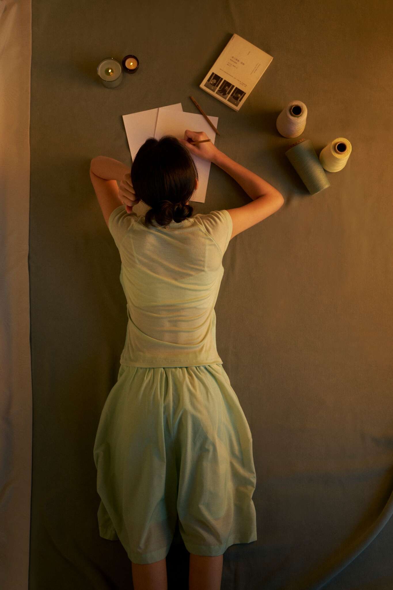 A person in light green clothes lies on their stomach, writing on paper. Nearby are two candles, a book, and spools of thread on a green surface, all softly lit.