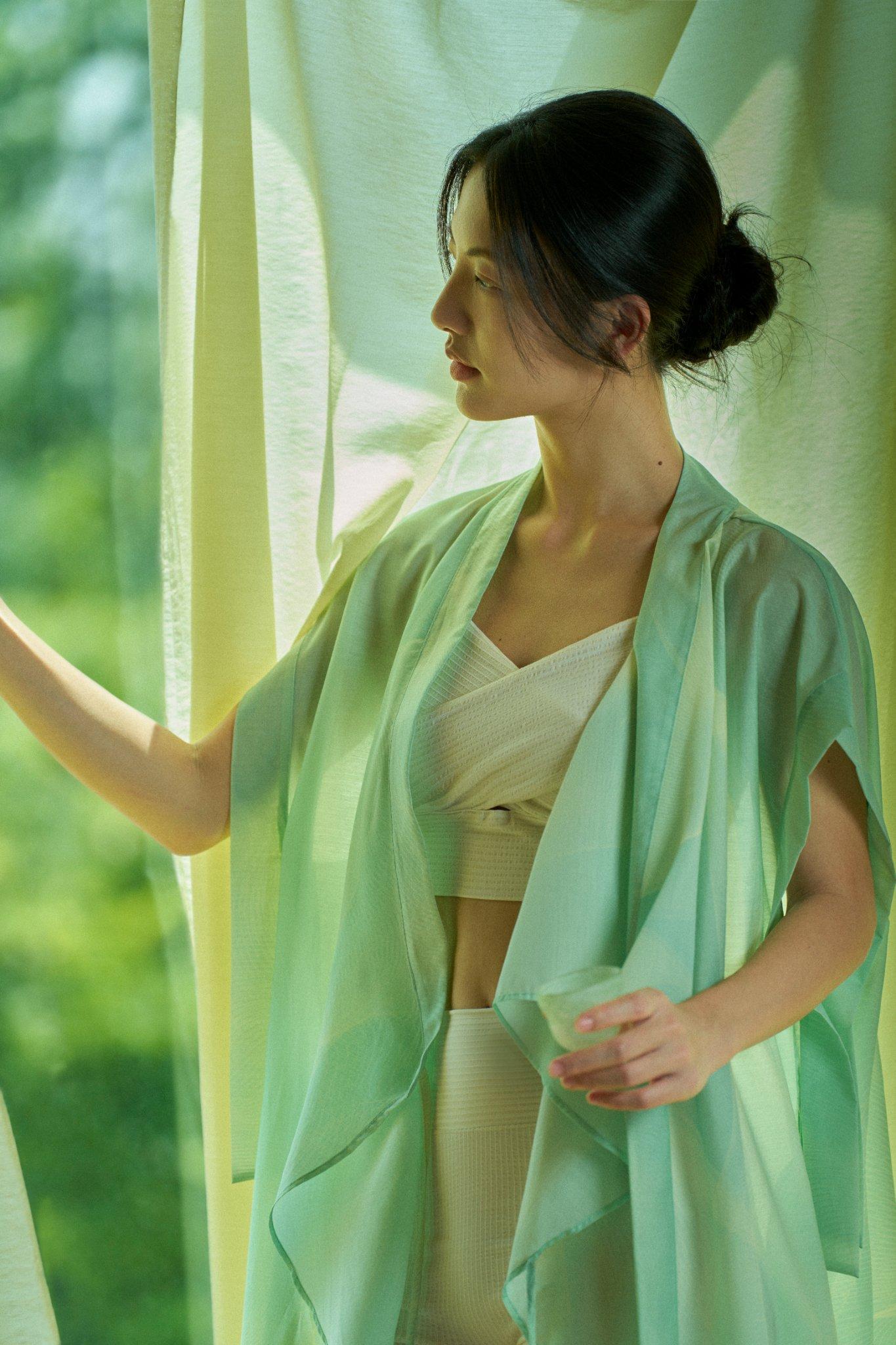A woman in light, airy clothing stands by a window with green curtains, holding a glass and looking outside. Sunlight creates a soft, serene atmosphere.