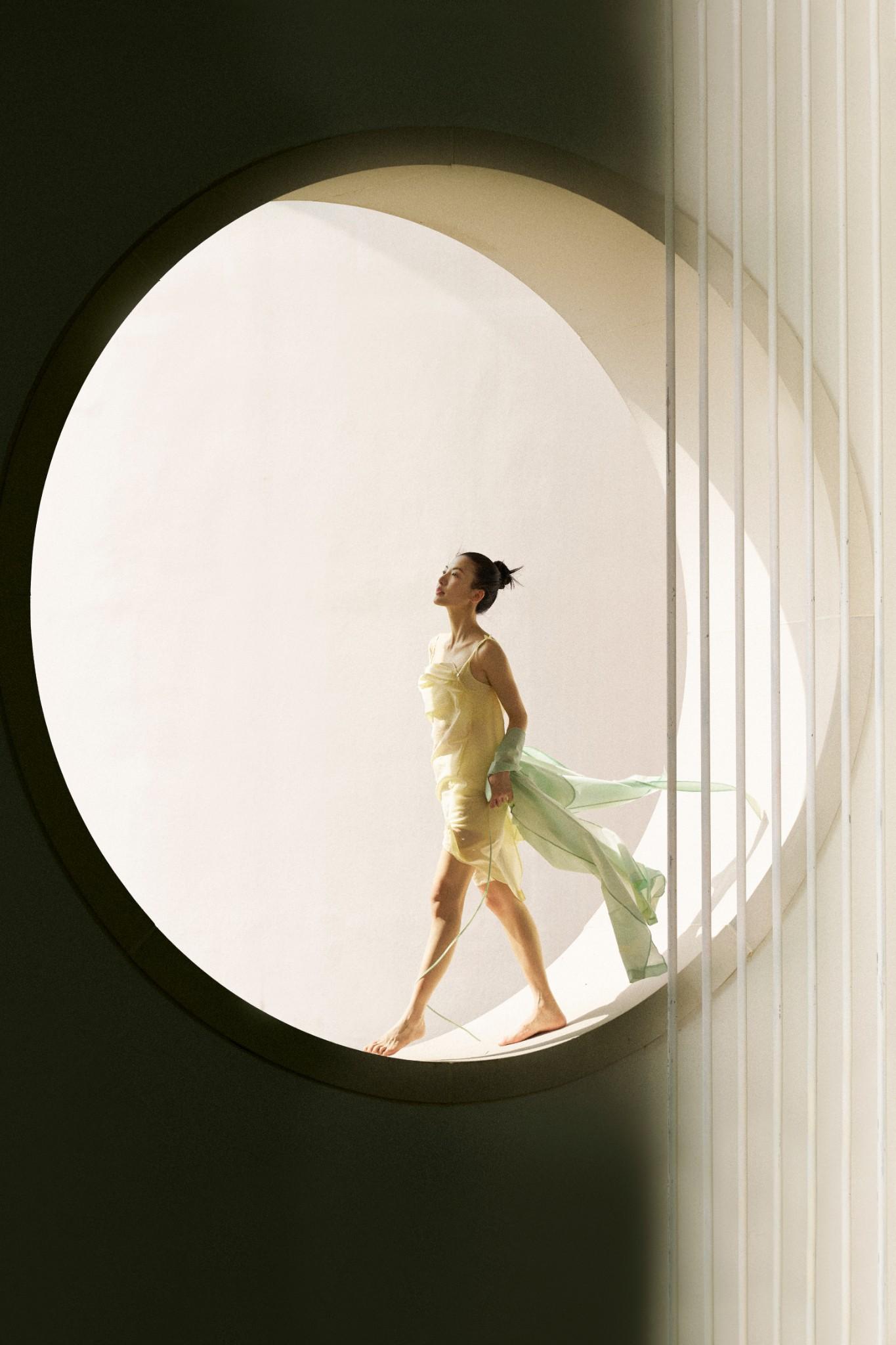 A woman in a flowing yellow dress walks barefoot inside a large circular architectural opening, sunlight illuminating her and casting soft shadows on the wall.