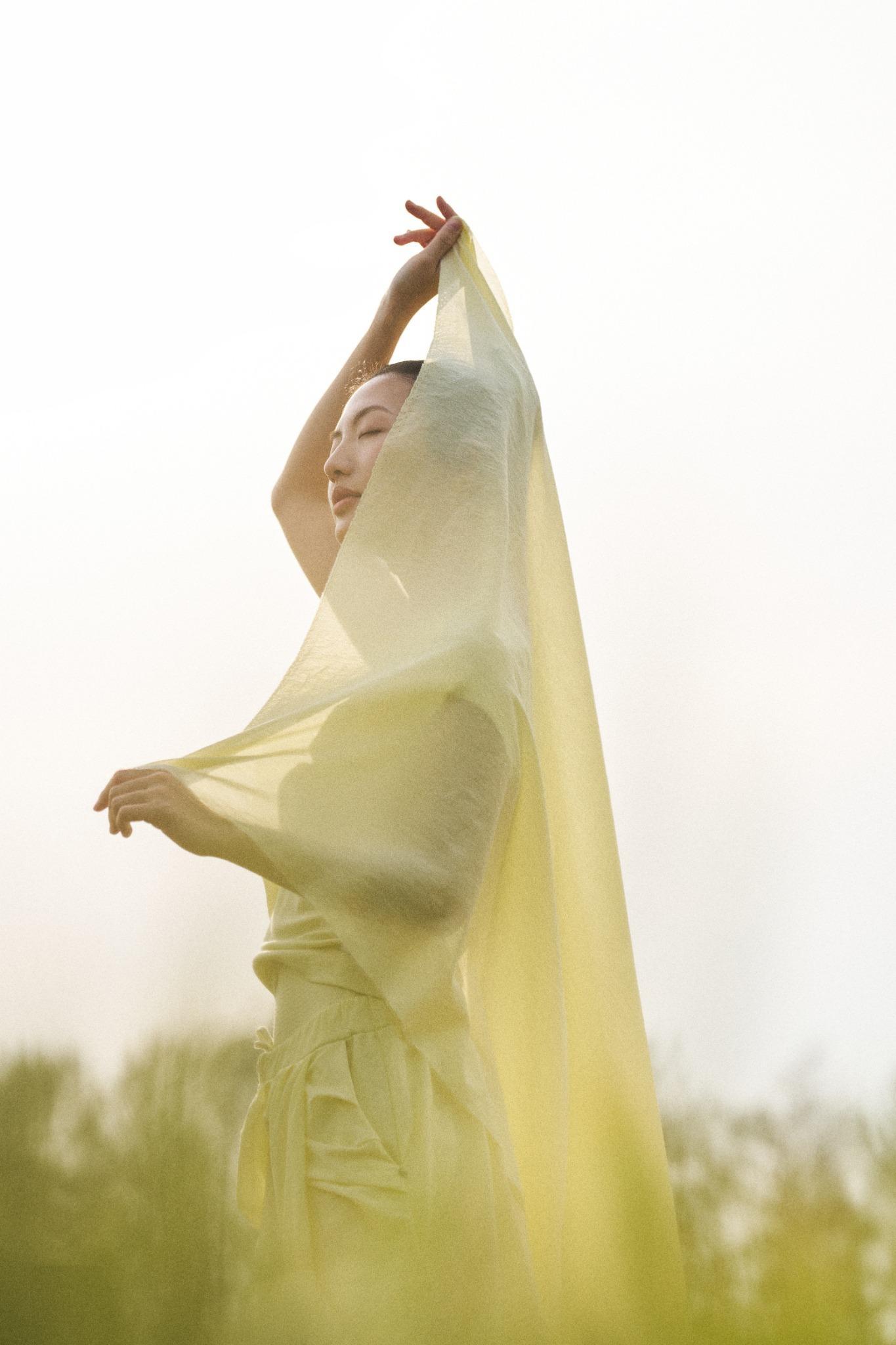 A person in a light green outfit poses outdoors, holding a sheer yellow fabric overhead, with soft sunlight and blurred trees in the background.